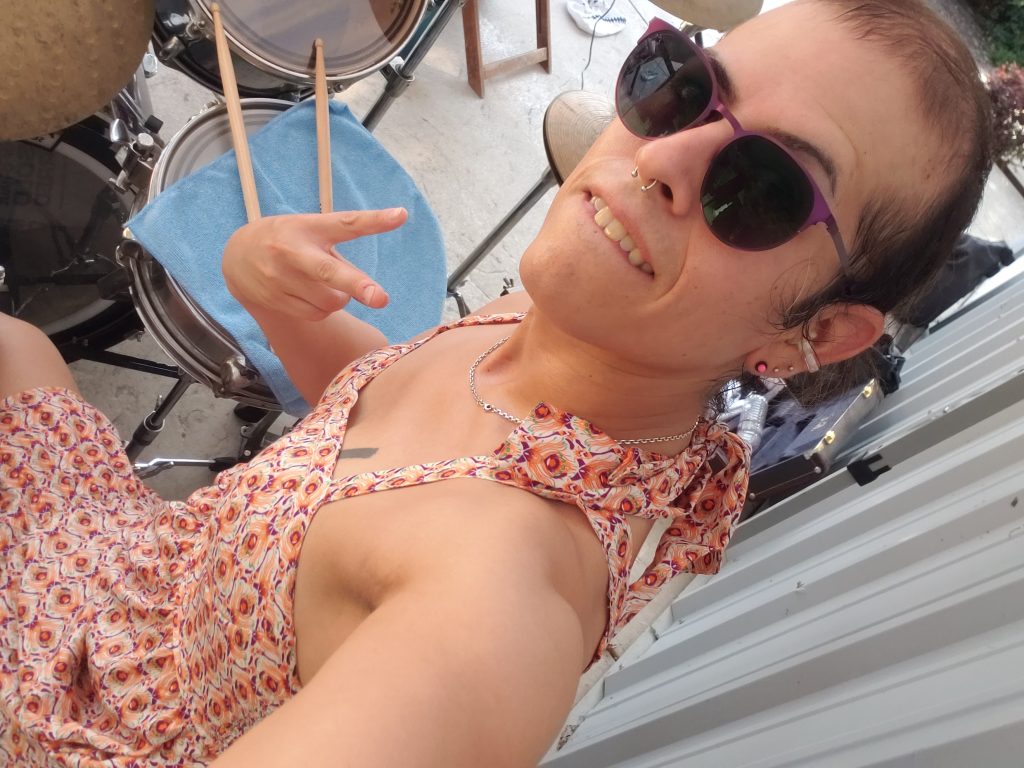 Vivi Dusk gritting her teeth at the camera while holding up devil horns over her drum set.