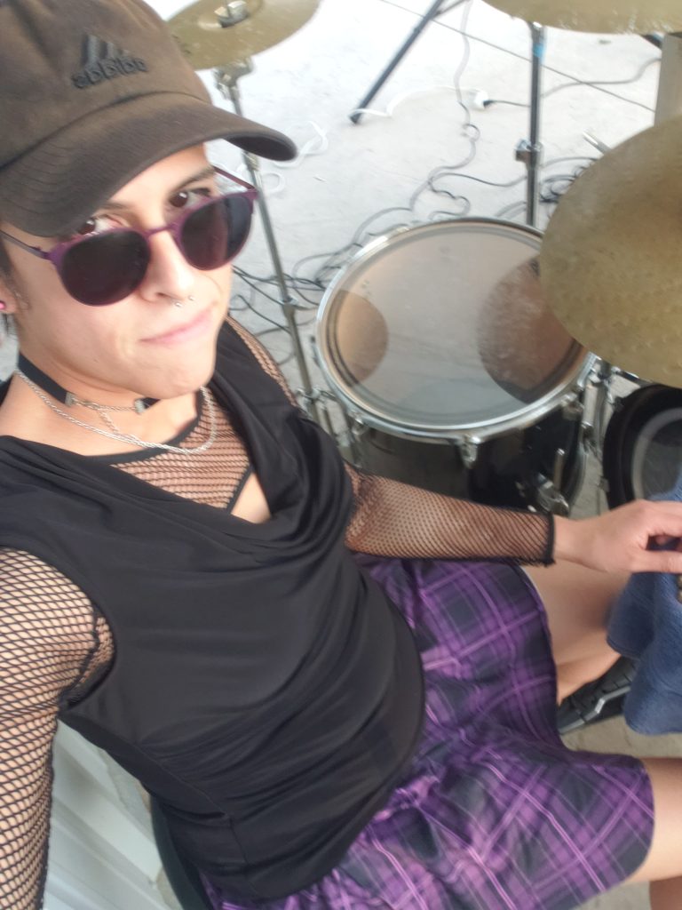 Vivi Dusk looking at the camera from above her sunglasses, sitting at her drumset.