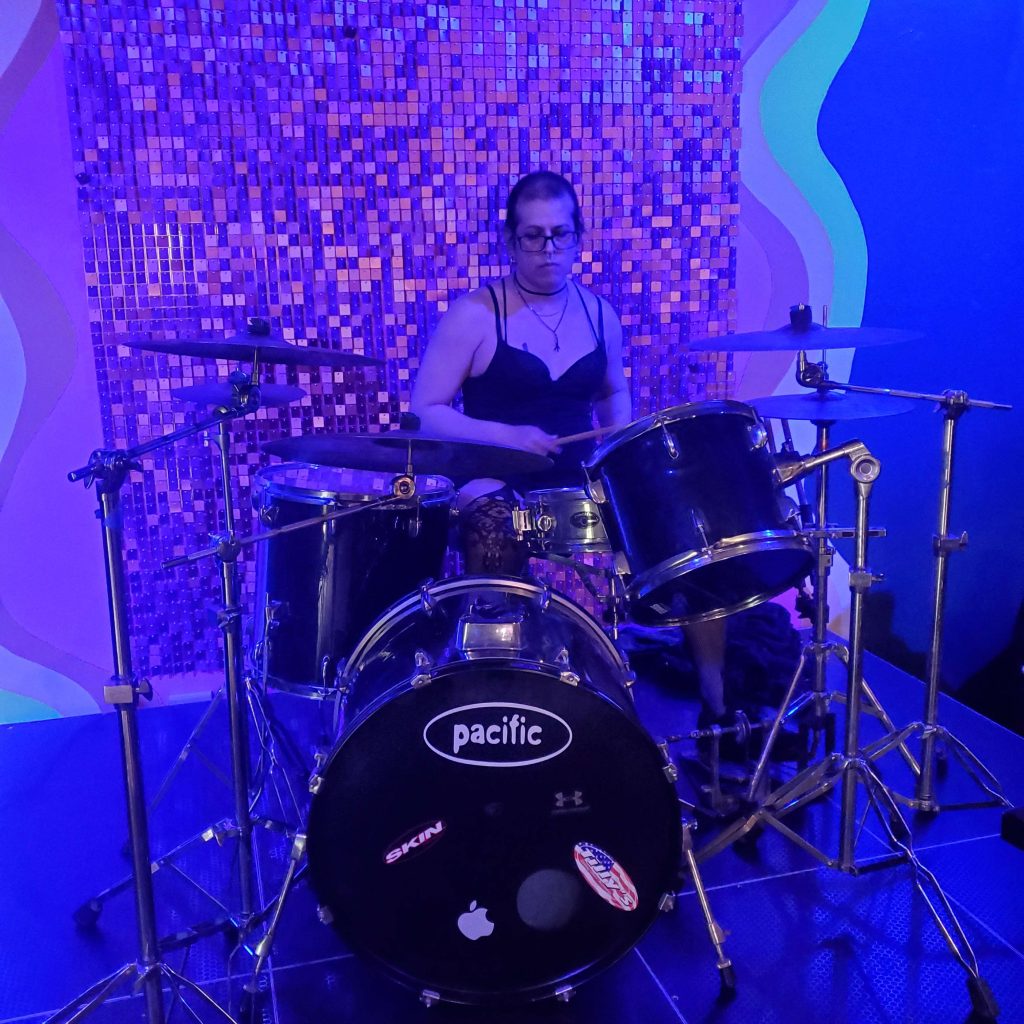 Vivi Dusk playing drums live at a club like venue.