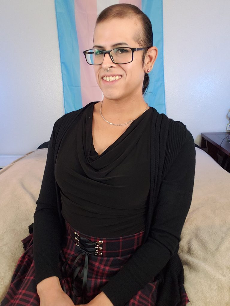 Vivi Dusk sitting at the edge of her bed and smiling in front of a trans pride flag on the wall. She's wearing a cute plaid red skirt wand black ruffled shirt.