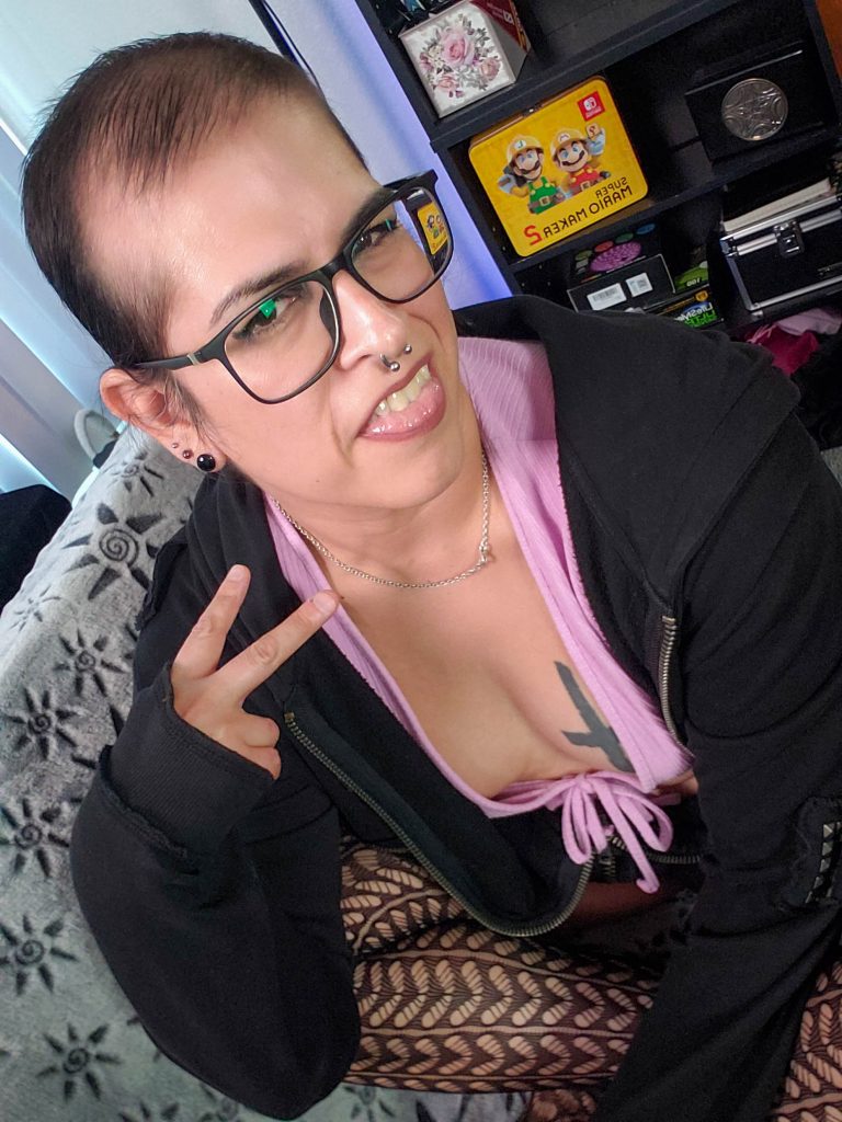 Soft goth selfie of Vivi Dusk wearing glasses, a black hoodie, pink top, fishnet tights, and piercings while posing with a peace sign.